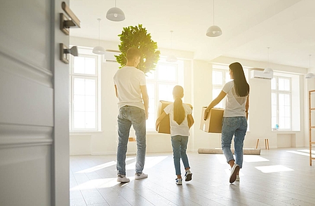 Familie trägt Umzugskartons und eine Pflanze in ein helles, modernes Wohnzimmer