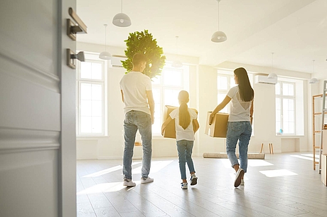 Familie trägt Umzugskartons und eine Pflanze in ein helles, modernes Wohnzimmer